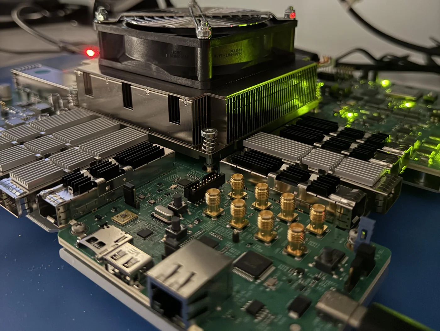 Versal FPGA prototyping board on a lab bench with heatsink fan, SFP cages, coaxial connectors and Ethernet, monitors behind